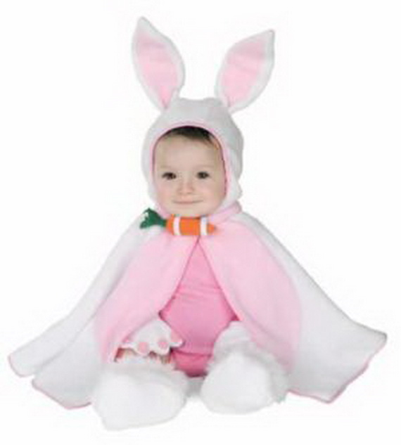 An infant girl sitting down while wearing a white and pink plush baby bunny rabbit Halloween costume cape with tall ears and a decorative carrot clasp.