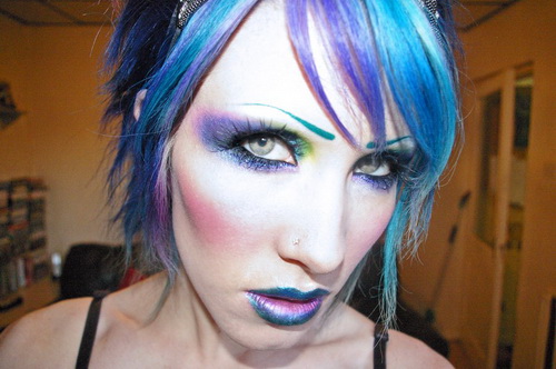 A woman displaying futuristic cyberpunk vampire makeup with purple and blue eyeshadow, a drawn-on teal eyebrow, metallic blue-rimmed ombre lips, and vibrant blue and purple hair.
