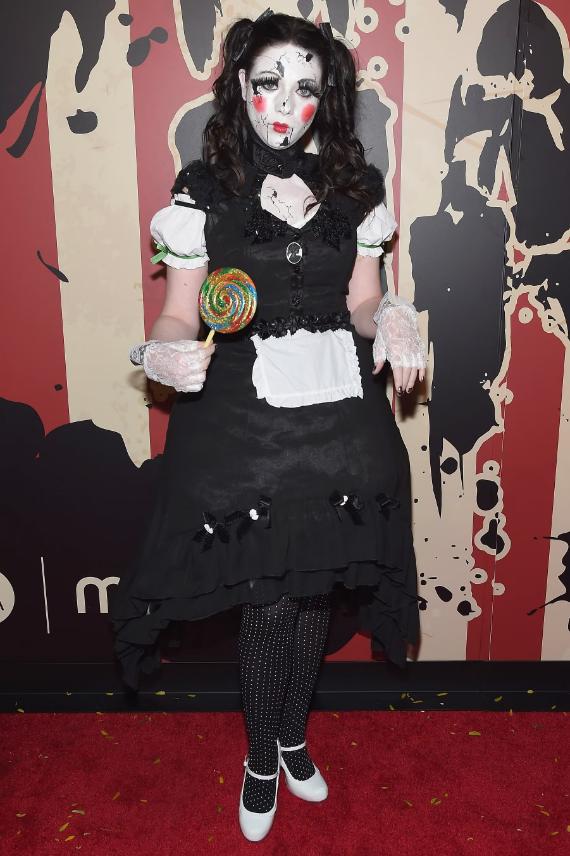 A woman dressed as a broken porcelain doll with cracks on her face holds a colorful lollipop on a red carpet.