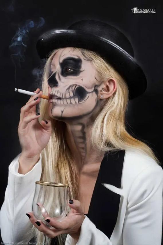 Woman with skeleton face paint, wearing a Chic-Halloween-Costumes-Skeleton , hat and jacket, smokes a cigarette while holding a glass.