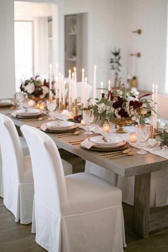 "Formal White and Gold Elegant Tablescape featuring tall lit white candles in a floral centerpiece on a dining table with white chairs. The setting uses gold utensils and decor, creating a Warm Formal Dinner Ambiance."