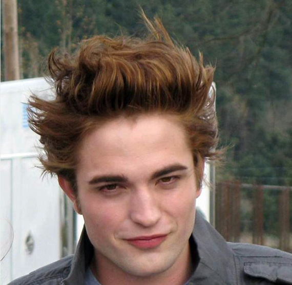 A person with voluminous, styled brown hair smirks slightly. They wear a gray collared shirt against a blurred outdoor background of trees.