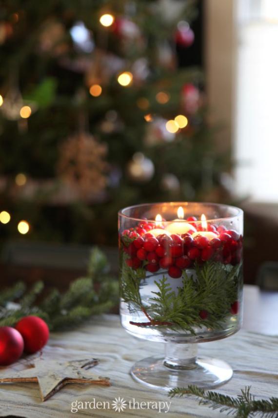 Christmas candles floating in a glass jar with red berries and greenery, glowing softly against blurred Christmas tree lights for a festive table centerpiece.