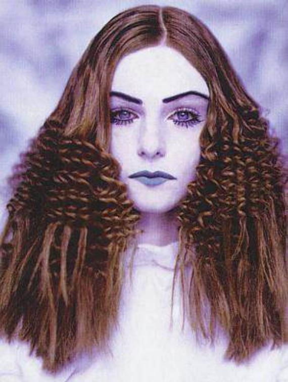 A woman with crimped brown hair and dramatic makeup stands against a soft, blurred background. Her expression is serious, creating a surreal, artistic tone.