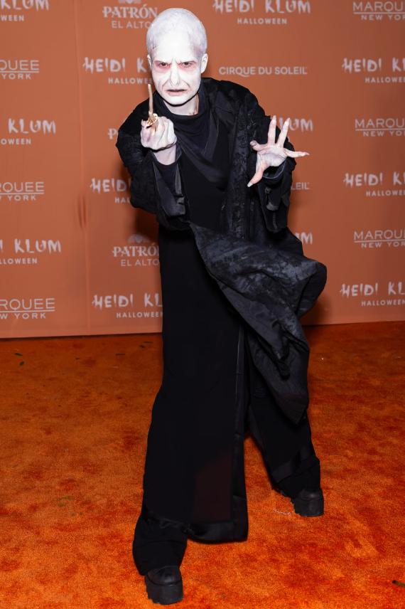 Person in Voldemort costume with white face paint and black robes stands on orange carpet.