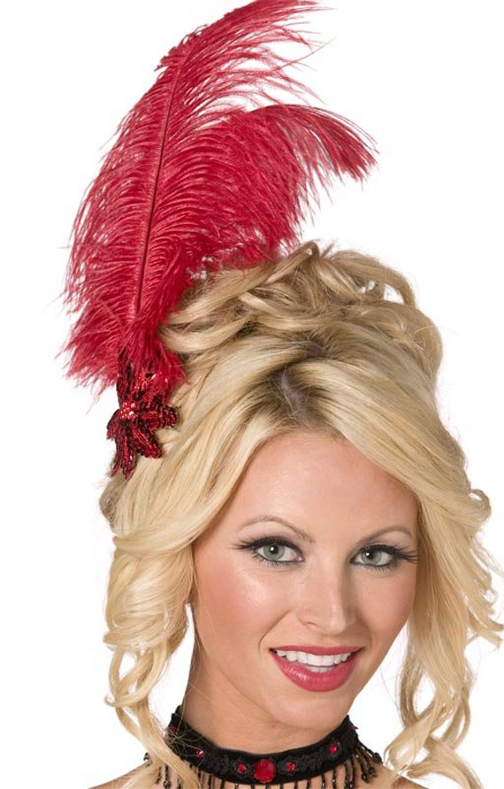 A woman with blonde curls wears a vintage-style updo adorned with a large red feather and decorative beaded detail. She smiles warmly, exuding a playful, elegant vibe.