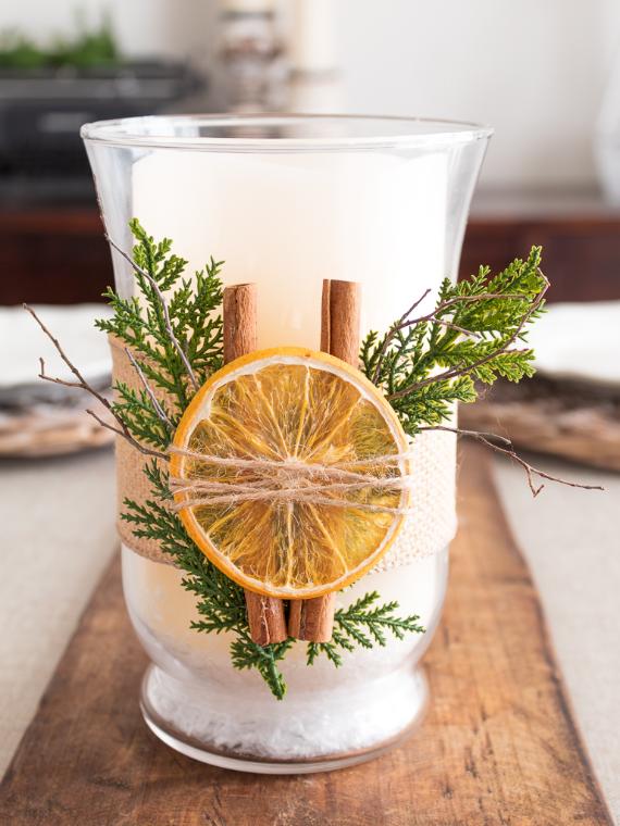"Rustic Aromatic Candle Decoration on a wooden table. A white candle in a clear glass holder is adorned with dried orange slices, cinnamon sticks, and evergreen sprigs, creating a Cozy Farmhouse Table Accent."