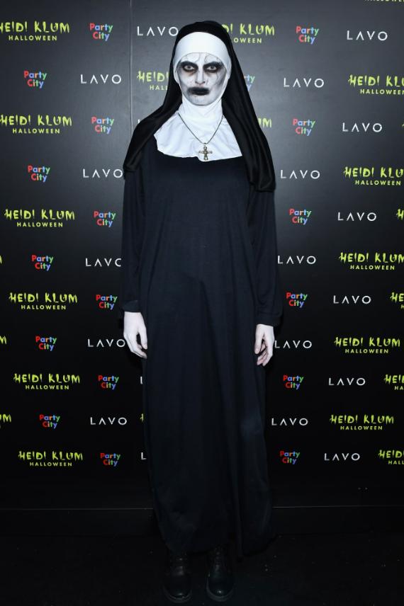 Person in a demonic nun costume with white face paint and a black habit stands against a black backdrop with "Heidi Klum Halloween" logos.