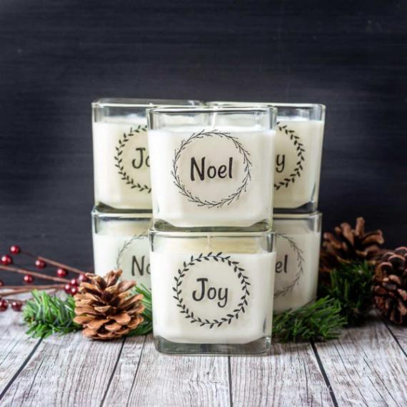"Close-up of a Noel and Joy Labeled Candle Vignette. Five stacked white candles in glass are nestled among Natural Pinecone and Evergreen Decor, providing a beautiful Cozy Holiday Spirit Accent for shelves or tables."