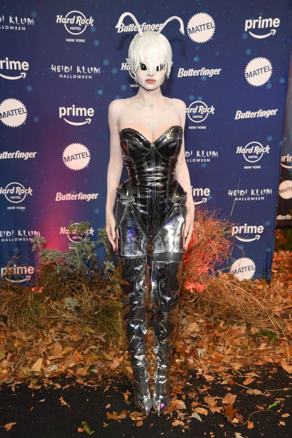 A woman in a shiny silver alien costume with white hair and antennae stands on a carpet of leaves in front of a backdrop with various sponsor logos.