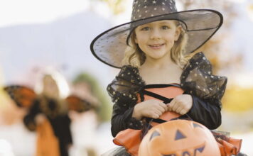The Halloween spirit of Trick-or-treating on Halloween Holiday A smiling young girl in a black and orange witch costume with a star-patterned hat holding a plastic pumpkin candy bucket outdoors.