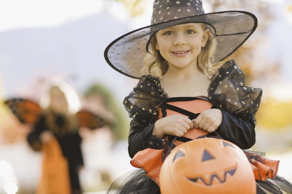 Young Girl in Cute Witch Costume Trick-or-Treating A smiling young girl in a black and orange witch costume with a star-patterned hat holding a plastic pumpkin candy bucket outdoors.