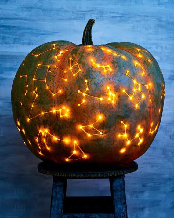 A large and Unique Jack-O’-Lantern Idea sits on a stool, carved with glowing star constellations. The warm, yellow-orange lights create a celestial, mystical ambiance.