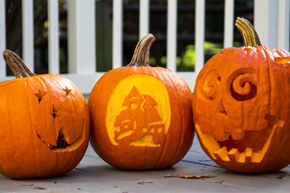 Three carved pumpkins on a porch; designs include a starry face, a haunted house, and a spiral-eyed grin, evoking a cheerful Halloween spirit.