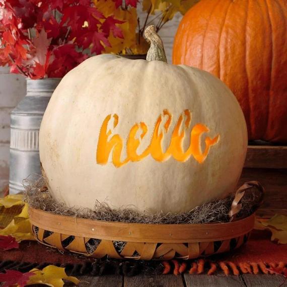 A Unique Jack-O’-Lantern Idea made of white pumpkin with "hello" carved in cursive sits in a basket on a wooden table. Autumn leaves are scattered around, with red foliage and another pumpkin in the background.
