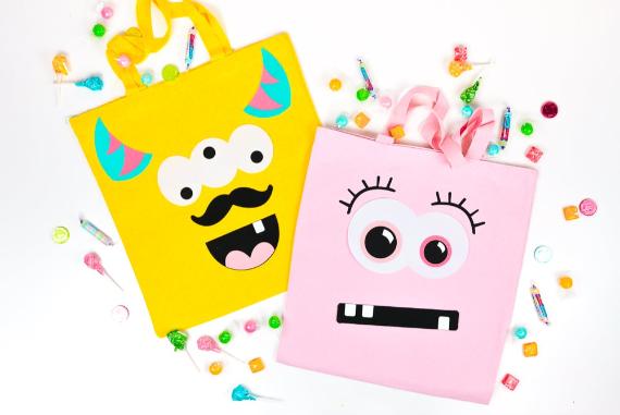 Eco-friendly Halloween bags for trick-or-treating feathering two colorful tote bags with playful monster faces; a yellow bag with horns and mustache, and a pink bag with big eyes. Scattered candy surrounds them.