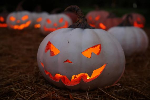 A White Scary Jack O’ Lanterns idea, pumpkins carved with glowing, sinister faces sit on straw. The dimly lit scene evokes a spooky Halloween atmosphere, with mischief in the air.