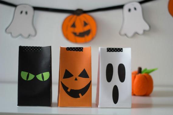 Three Halloween-themed paper bags are on a table: a black bag with green eyes, an orange bag with a jack-o'-lantern face, and a white bag with a ghost face. A string of ghost and pumpkin decorations is hung in the background, creating a festive atmosphere.
