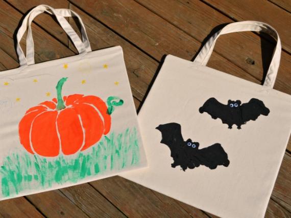 Two canvas tote bags on a wooden surface. One bag has a painted orange pumpkin on green grass with stars. The other shows two black bats. Halloween theme.