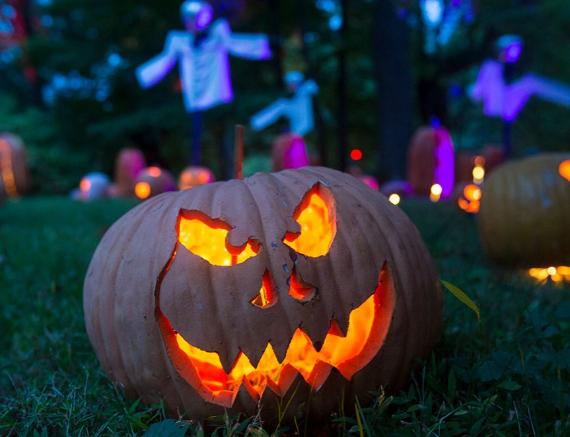 A glowing jack-o'-lantern with a menacing grin sits on grass. The background features purple-lit scarecrows and pumpkins, creating a spooky Halloween scene.