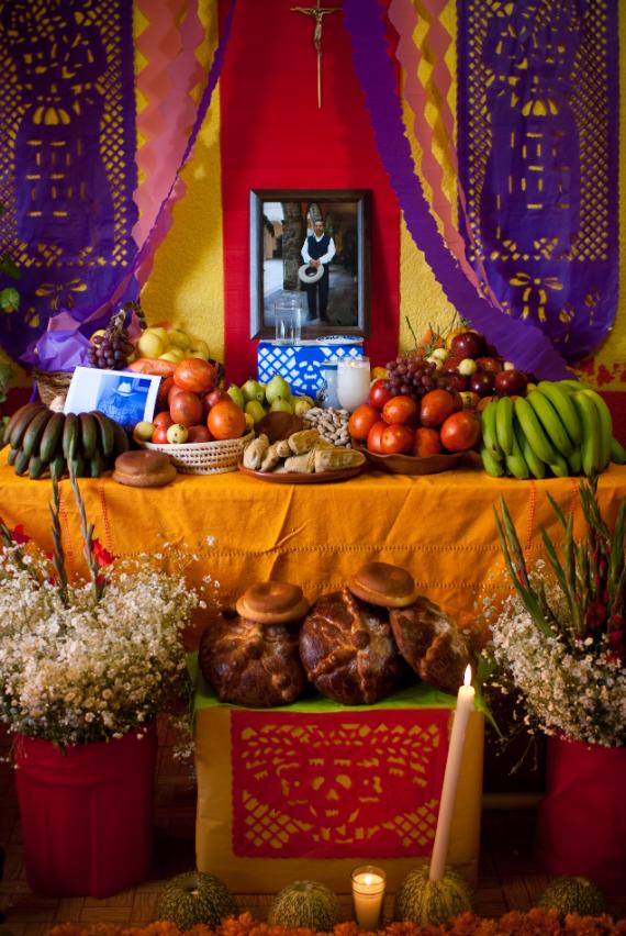 A traditional multi-tiered Day of the Dead altar featuring a framed family photo, purple papel picado with skull cutouts, pan de muerto, fresh fruit, candles, and flowers.