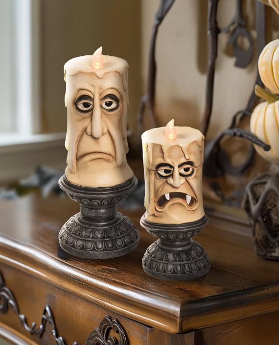 Two sculpted cream-colored Halloween LED candles with expressive monster faces on dark gothic pedestals.