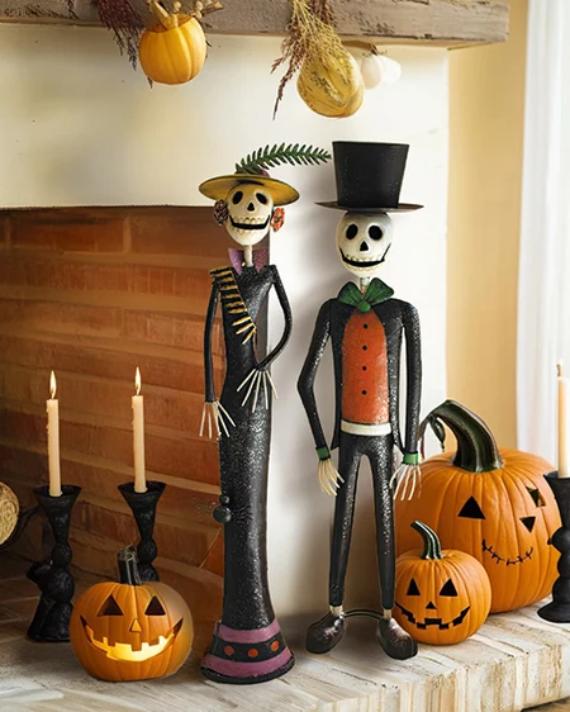 Two tall, painted metal Day of the Dead skeleton figures representing a Catrina in a yellow sun hat and a Catrín in a black top hat standing on a fireplace mantel.