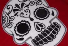 Day of the dead craft skull designs A large, detailed white felt patch embroidered with black thread in a traditional sugar skull design, displayed on a solid red fabric surface.