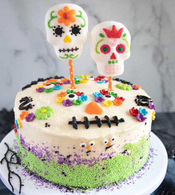A festive Day of the Dead cake coated in white frosting, green and purple sprinkles, and colorful frosting flowers, topped with two large marshmallow sugar skull pops for All Souls' Day.