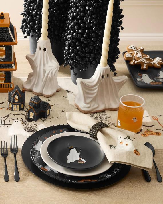 A festive Halloween table setting with white ceramic ghost candle holders, ghost-printed plates, napkins, and miniature haunted houses.