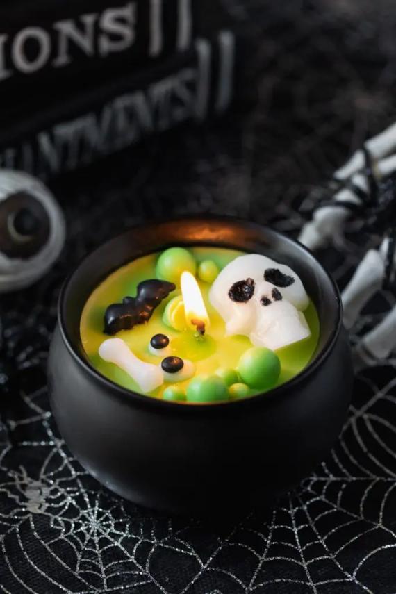 A lit Halloween cauldron candle filled with bubbling green wax, a white wax skull, a black bat, and bones on a spiderweb tablecloth.