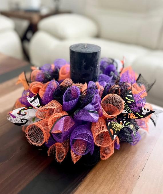 A thick black pillar candle centered inside an orange, purple, and black deco mesh holiday wreath ring on a wooden table.