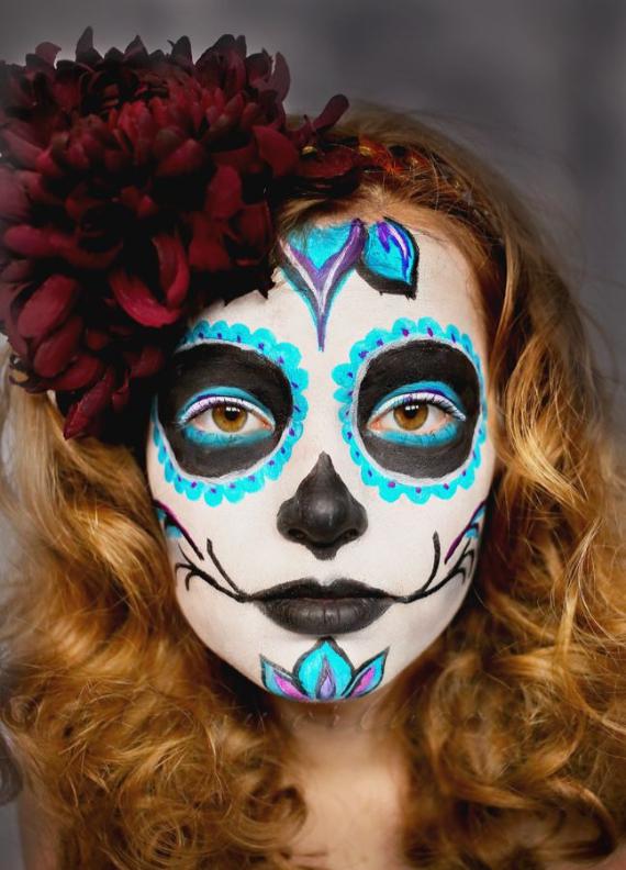 A young girl with blonde curly hair wearing traditional Day of the Dead face paint featuring a white base, black eye sockets with turquoise scalloped edges, and a dark red floral hair crown. 