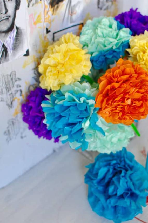 A bouquet of handmade tissue paper flowers in bright orange, yellow, turquoise, mint green, and purple arranged next to an ancestor's sketch on a Day of the Dead altar.