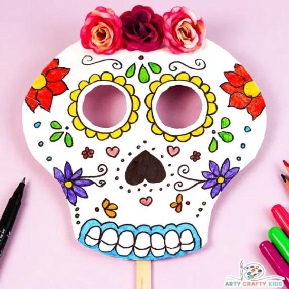 A hand-colored printable paper sugar skull mask on a wooden stick decorated with red, purple, and yellow floral designs and topped with artificial pink and red roses for Day of the Dead activities.