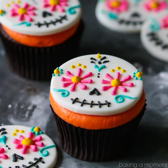 Close-up of a chocolate cupcake topped with bright orange frosting and a smooth white fondant disk decorated with a pink, yellow, and blue royal icing sugar skull pattern for Day of the Dead celebrations.