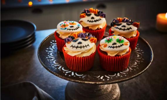 Five chocolate cupcakes in red wrappers on a vintage metal stand decorated with white fondant sugar skull faces and colorful frosting flowers for Day of the Dead activities.