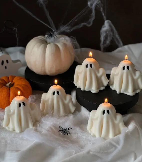 Five lit miniature white ghost candles arranged on a table with white and orange pumpkins, faux spiderwebs, and plastic spiders.