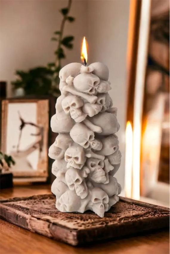 A tall, sculpted grey wax candle shaped like a stacked tower of human skulls and bones, lit on a rustic wooden stand.
