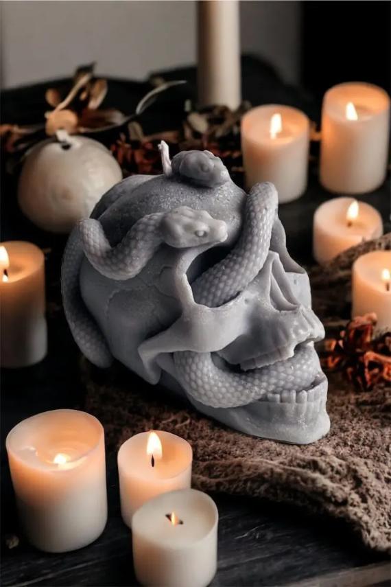 A large grey wax skull candle sculpted with slithering snakes surrounded by glowing white votive candles on a dark table.