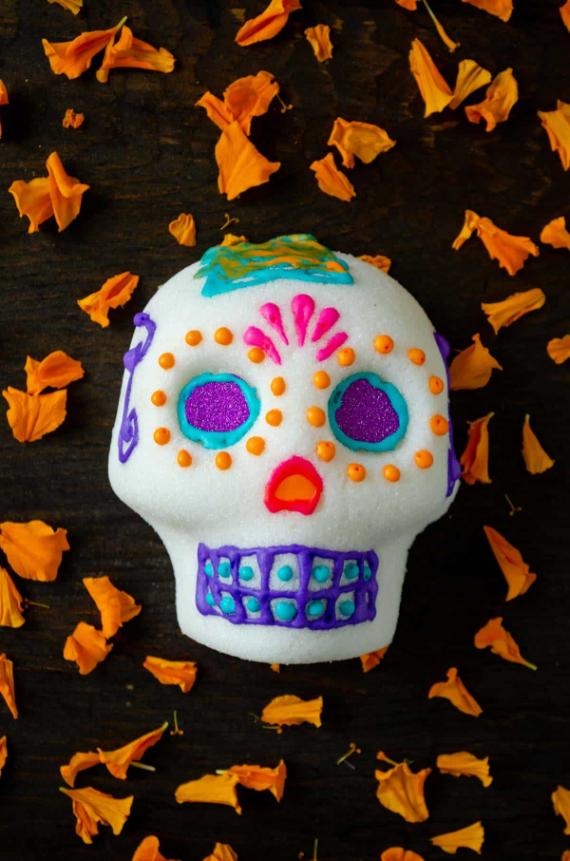 An authentic molded sugar skull with purple glitter eyes, pink and orange icing details, resting on a dark wood background surrounded by orange marigold petals.