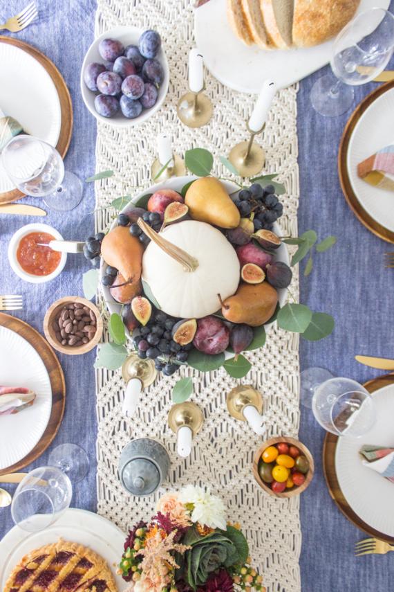 Festive dining table with a centerpiece of a white pumpkin, pears, figs, and grapes. Surrounded by elegant tableware, bread, and fall colors on a woven runner.