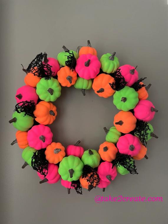 A vibrant wreath of Non-traditional fall color palette of small pumpkins in neon pink, orange, and green. Black mesh accents add texture against the gray background, creating a festive and lively tone.