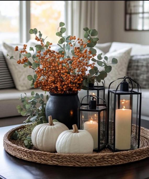 An elegant blue fall decorations centerpiece featuring a woven tray with white pumpkins, orange berries, eucalyptus, and lit candles in black lanterns.