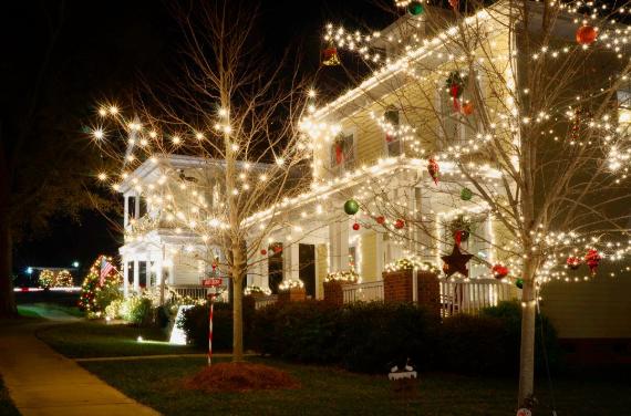 Two houses adorned with festive Christmas lights at night. Warm white lights cover trees and eaves, with red and green ornaments adding color.