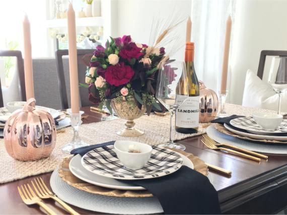 Festive dining table with elegant gold and black plaid settings, copper pumpkins, tall candles, vibrant floral centerpiece, and a wine bottle.