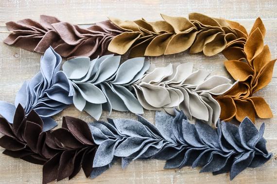 Fabric leaves in autumn shades of brown, gray, and beige are artfully arranged in a horizontal pattern on a textured wooden surface, evoking a warm, cozy feel.