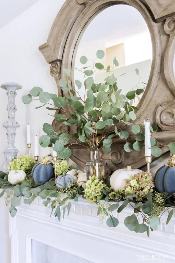 Blue fall decorations for mantel with rustic mirror, decorated with eucalyptus branches, white and blue pumpkins, hydrangeas, and tall candlesticks. Cozy, autumnal feel.