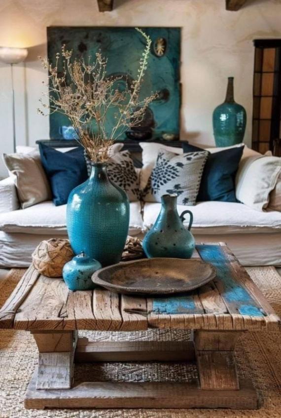 A cozy living room with a rustic wooden table adorned with teal vases and dried flowers. A white couch with patterned and blue cushions sits behind.