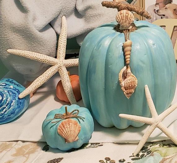 A coastal-themed decor featuring blue pumpkins adorned with seashells and starfish, set against a soft backdrop. The tone is beach-inspired and serene.